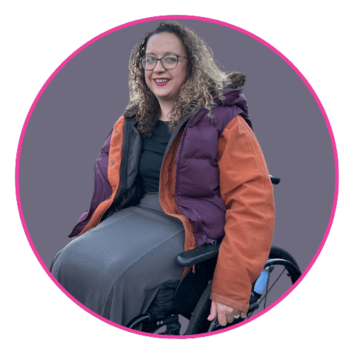 Carrie-Ann seated in a wheelchair, smiling and wearing a purple and orange winter jacket over a black top and long grey skirt.
