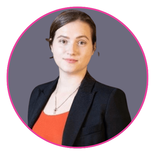 Heloise wearing a black blazer over an orange top, with brown hair tied back and a small pendant necklace.