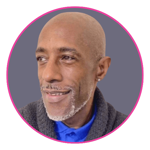 Carl with a neatly groomed grey beard and bald head, wearing a dark textured sweater over a blue collared shirt and a small earring.