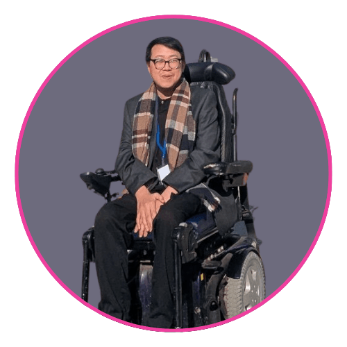 James Lee seated in a motorised wheelchair against a grey background, wearing a dark blazer and checkered scarf.